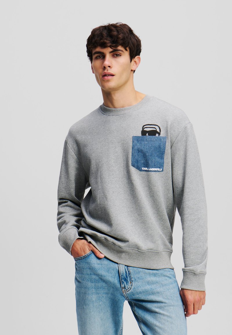 Felpa grigia con tasca in denim e design grafico. Vestibilità comoda, colletto rotondo e polsini a coste. Indossata con jeans azzurri chiari.