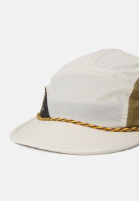 Casquette avec un avant beige, des côtés vert olive et un panneau en maille noir. Accentuée par une bande tissée colorée le long de la visière. Matériau léger.