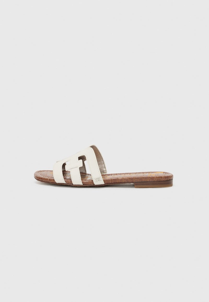 Flat glidesandal med en strukturert brun såle og en off-white skinnoverdel med to brede stropper og et utskjært design.
