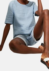 Ljusblå, t-shirt med kort ärm och rund halsringning, kombinerad med denimshorts. Bärs med vita slidesandaler och visar en detaljerad tatuering på benet.