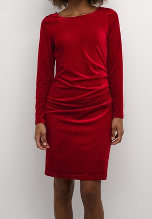 Frau trägt ein figurbetontes, langärmliges rotes Samtkleid mit gerafften Details an der Taille, steht vor einem schlichten weißen Hintergrund.