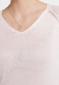 Pull en maille rose clair à col en V, présentant une texture fine et un tissage aéré, accompagné d'un collier délicat en pendentif doré.