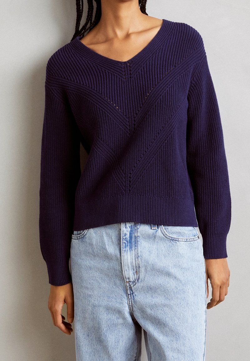 Navy blue knitted sweater with a V-neck, featuring textured chevron patterns and ribbed cuffs. Paired with light blue, high-waisted jeans.