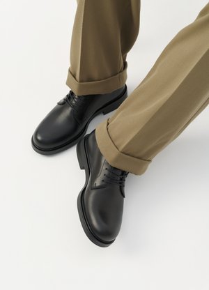 Chaussures habillées en cuir noir avec lacets portées avec un pantalon kaki retroussé sur fond blanc.