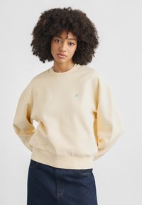 Φούτερ σε ανοιχτό μπεζ βαμβάκι με μακριά, χαλαρά μανίκια, ριμπ λαιμόκοψη και στρίφωμα. Διαθέτει μικρό λογότυπο στο στήθος.