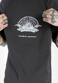 Black t-shirt featuring a silver graphic of a decorative banner with "MULTIPLY APPAREL" and the number "10" above "multiply apparel."