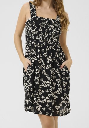 Femme portant une robe noire sans manches avec un imprimé floral blanc et des poches, debout devant un fond clair uni.