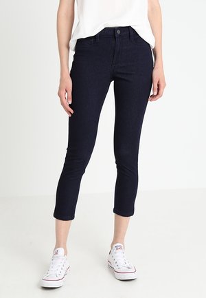 Frau trägt dunkelblaue, kurze Skinny-Jeans, weiße Freizeitsneaker und ein lockeres weißes Oberteil, steht vor einem schlichten hellen Hintergrund.