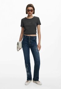 T-shirt grigia corta, jeans di denim blu con ricami floreali e espadrillas di colore chiaro. Accessori: borsa bianca, occhiali da sole scuri.