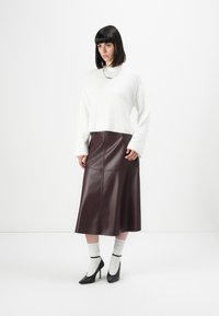 Vrouw in een witte fluffy trui, bordeaux leren midi-rok, witte sokken en zwarte hoge hakken, staand op een witte achtergrond.