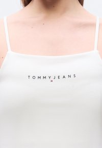 Λευκό τοπ με λεπτές τιράντες, με το κείμενο "TOMMY JEANS" και μικρό λογότυπο Tommy Hilfiger στο κέντρο του στήθους.