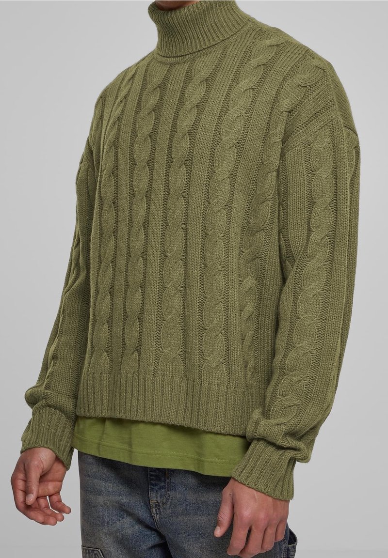 Pull à col roulé en tricot torsadé vert olive avec poignets et ourlet côtelés, porté sur une chemise vert clair, affiché sur un fond neutre.