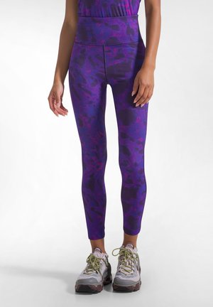 Kvinde iført lilla mønstrede leggings med høj talje og grå sportssko med grønne snørebånd, stående på en hvid baggrund.