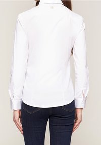 Vit skjorta med hög krage, långa ärmar och slitsar vid ärmsluten. Minimalistisk design med en liten logotyp nära halsringningen.