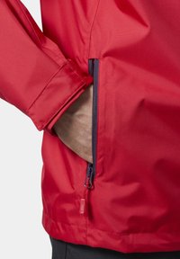 Czerwona kurtka z zamkiem błyskawicznym i teksturowanym materiałem. Zamek ma kontrastujący, fioletowy uchwyt. Zbliżenie na rękaw.