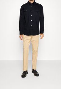 Chemise noire à manches longues avec deux poches poitrine, devant boutonné, associée à un pantalon beige et des chaussures noires, se tenant sur un fond uni.