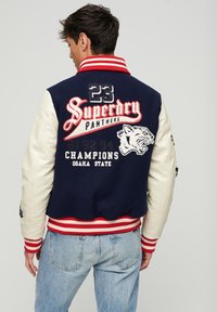 Chaqueta varsity azul marino y crema con acentos rojos. Presenta texto bordado, números y un diseño de tigre en la parte posterior. Puños y dobladillo acanalados.