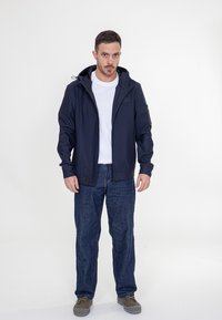 Granatowa kurtka z kapturem, zamkiem błyskawicznym z przodu i elastycznymi mankietami, zestawiona z ciemnymi dżinsami. Posiada logo na piersi oraz teksturę materiału.