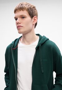 Grøn zip-up hoodie med en struktureret hætte, der har en hvid t-shirt underneath. Lille "HUGO" logo på venstre bryst og rød accent på lynlåsen.