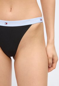 Γυναίκα που φοράει μαύρα εσώρουχα υψηλής κοπής με λευκή ζώνη μέσης με τη σήμανση "Tommy Hilfiger" και μικρό λογότυπο στο πλάι.