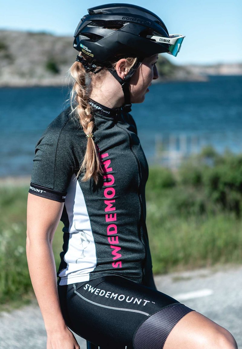 Maglia da ciclismo in bianco e nero con logo rosa, dotata di zip, abbinata a pantaloni corti neri. Casco nero e occhiali da sole. Ambientazione all'aperto.