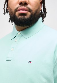 Άνδρας με γενειάδα που φοράει μπλούζα πόλο σε μέντα πράσινο χρώμα με μικρό ορθογώνιο λογότυπο σε κόκκινο, λευκό και μπλε στο στήθος, περικο μμένη φωτογραφία στο λαιμό και στους ώμους.