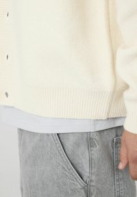 Cardigan en tricot crème avec un ourlet côtelé et une fermeture à bouton, associé à un T-shirt gris clair et un pantalon gris avec des poches latérales.