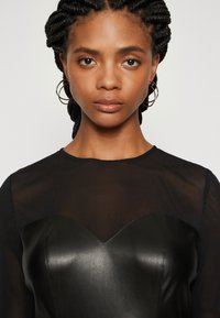 Corset din piele neagră cu decolteu în formă de inimă, purtat cu un top transparent negru. Părul împletit și cerceii tip cerc completează look-ul.