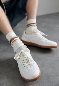 Baskets blanches en cuir avec une languette gris texturée, des lacets crème et une semelle en caoutchouc brun clair. Portées avec des chaussettes à rayures crème et beige côtelées.