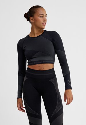 Kobieta ubrana w czarny bezszwowy krótki top z długim rękawem i legginsy z wysokim stanem z geometrycznymi wzorami, stojąca na tle jednolitego szarego tła.