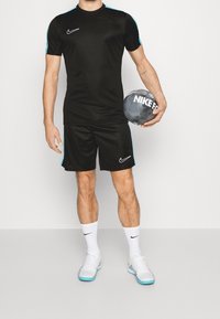 Schwarzes Sportshirt und Shorts mit türkisfarbenen Akzenten; leichtes Material, enganliegendes Design. Hält einen schwarzen Fußball mit "NIKE FC"-Markierung.
