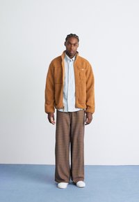Man wearing a brown fleece jacket over a light blue shirt, wide-legged plaid pants, and white shoes, standing on blue floor against white wall.