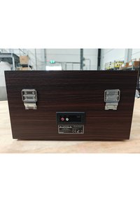 Panel trasero de dispositivo electrónico de madera con dos pestillos de metal, puertos de salida de audio RCA, entrada de alimentación y etiquetas de advertencia sobre una mesa de trabajo.