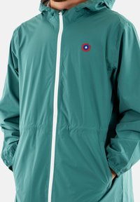 Veste coupe-vent teal avec une fermeture éclair blanche, arborant un logo circulaire brodé sur la poitrine et des poignets élastiques ; légère et résistante à l'eau.
