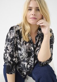 Blusa nera a fiori con motivi grigio chiaro e bianchi, maniche arricciate e chiusura abbottonata. Indossata con jeans blu scuro.