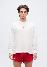 Νέος άνδρας με σκούρα μαλλιά που φορά λευκό μακρυμάνικο πουκάμισο και κόκκινα αθλητικά σορτς με μικρά κόκκινα λογότυπα, στέκεται μπροστά σε απλό φόντο.