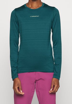 Persoon draagt een teal longsleeve met het "La Sportiva"-logo en magentakleurige broek, staand tegen een effen lichte achtergrond.