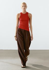 Rød ærmeløs top, tætsiddende design; brune stribede brede bukser; flade sorte sandaler. Model går på et neutralt gulv mod en ensfarvet baggrund.
