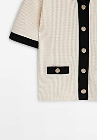 Veste beige texturée à manches courtes avec bordure noire, boutons dorés sur le devant et sur une poche bordée de noir.
