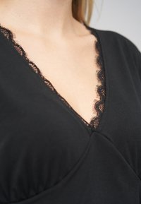 Schwarzes Top mit V-Ausschnitt und gewelltem Spitzenbesatz, gefertigt aus weichem Stoff mit drapiertem Design, zeigt eine glatte Textur.