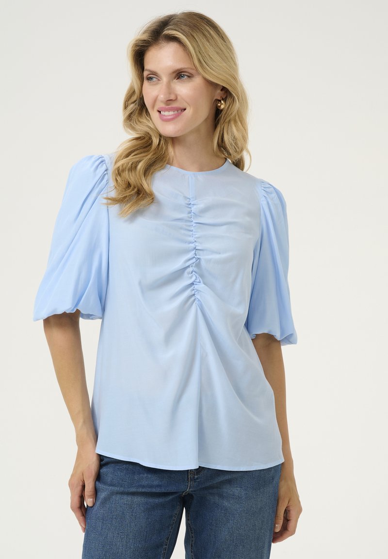 Gaiši zila blūze ar īsām pūkainām piedurknēm, ar savilktu priekšpusi, gludu tekstūru un apaļu izgriezumu, saskaņota ar džinsu biksēm.