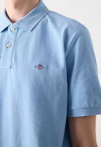 Homme portant un polo à manches courtes bleu clair avec col et deux boutons, arborant un petit emblème brodé sur le côté gauche de la poitrine.