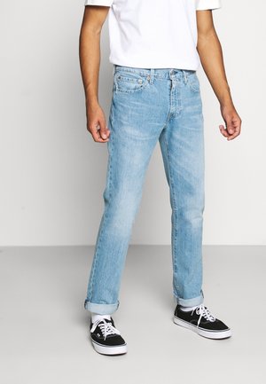 Jean en denim bleu clair à coupe droite, avec des ourlets retroussés et un design classique à cinq poches. Porté avec des baskets noires et un t-shirt blanc.