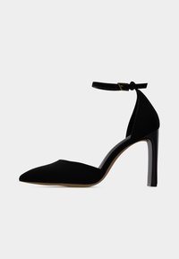 Μαύρο γυναικείο παπούτσι με ψηλό τακούνι και μυτερή μύτη, με λουρί στο αστράγαλο και χρυσή αγκράφα, σε λευκό φόντο, πλευρική όψη.