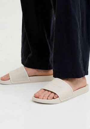 Lábak bézs csúszásgátló szandálban, széles pántokkal és fekete nadrágban, világos felületen állva.