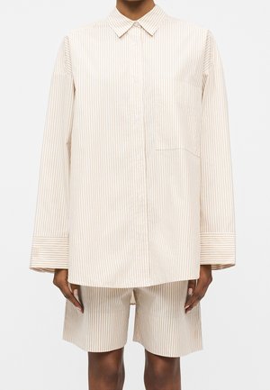 Person iført beige og hvide lodrette stribede oversized skjorte med brystlomme og matchende knælange shorts.