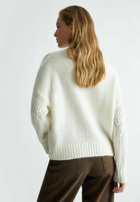 Maglione bianco a maglia con vestibilità ampia, maniche testurizzate con dettaglio a maglia aperta e orlo a coste. Abbinato a pantaloni marroni.