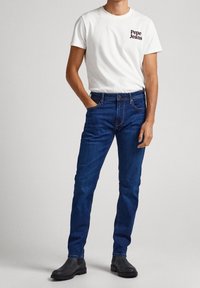 White short-sleeve t-shirt with "Pepe Jeans" logo, paired with dark blue fitted jeans and black ankle boots. Minimalist design, casual style.