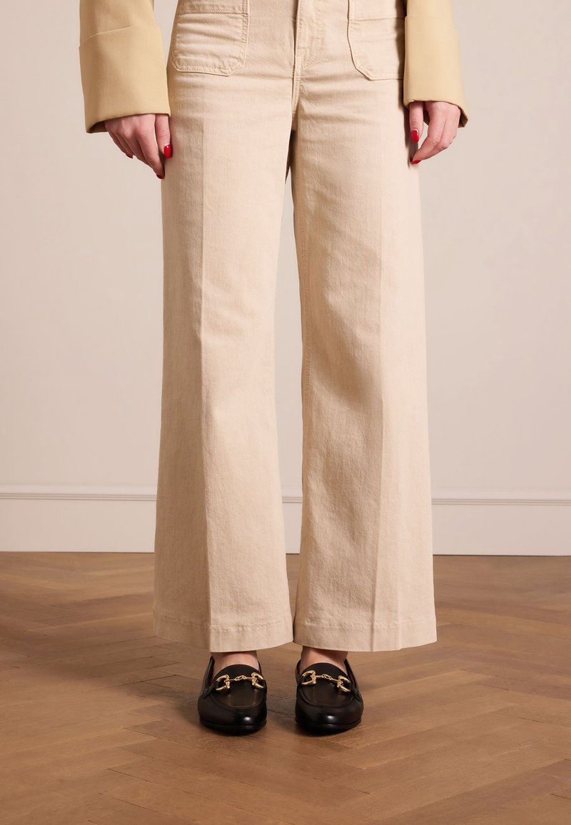 Femme portant un pantalon beige à jambes larges et des mocassins noirs avec des détails en chaîne dorée, debout sur un sol en bois contre un mur uni.