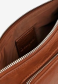 Interno di una borsa in pelle marrone con una superficie liscia, caratterizzato da una cerniera in argento e un'etichetta in pelle marchiata posizionata all'interno.